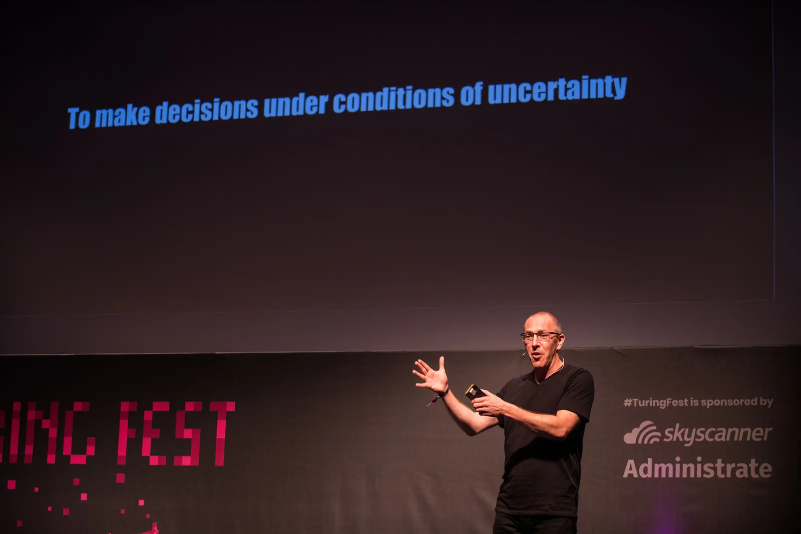 Fred Destin gesturing on stage at Turing Fest, slide behind him reads 'To make decisions under conditions of uncertainty'