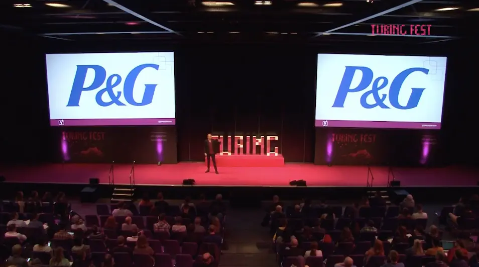 Jono Alderson on stage at Turing Fest; twin screens display the P&G logo